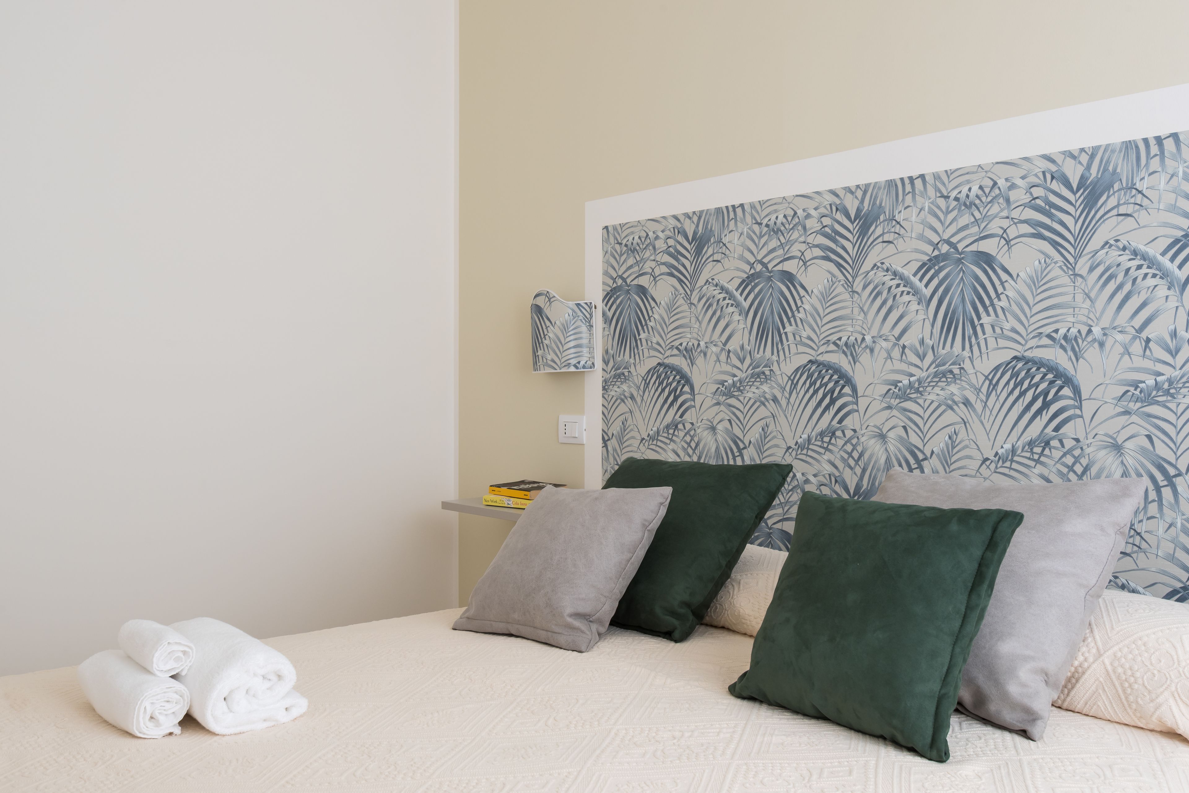 Un lit bien fait, avec des serviettes blanches pliées en rouleau, une table de chevet avec des livres et un mur orné d’un motif de feuilles tropicales.
