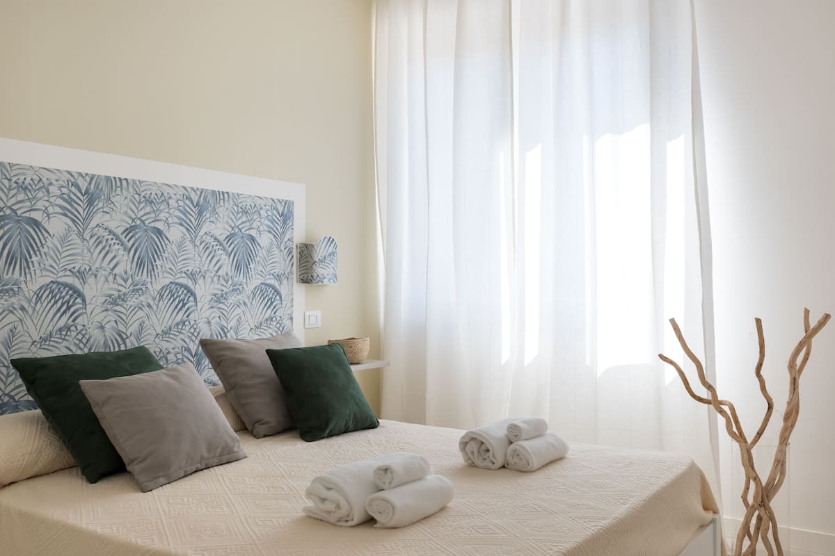Un lit bien fait, avec des serviettes pliées en rouleaux, une tête de lit ornée d’un motif de feuilles et une fenêtre avec des rideaux légers.