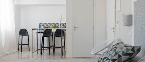 A modern hotel room with a bed, a desk with chairs, a small kitchen area, and a door.