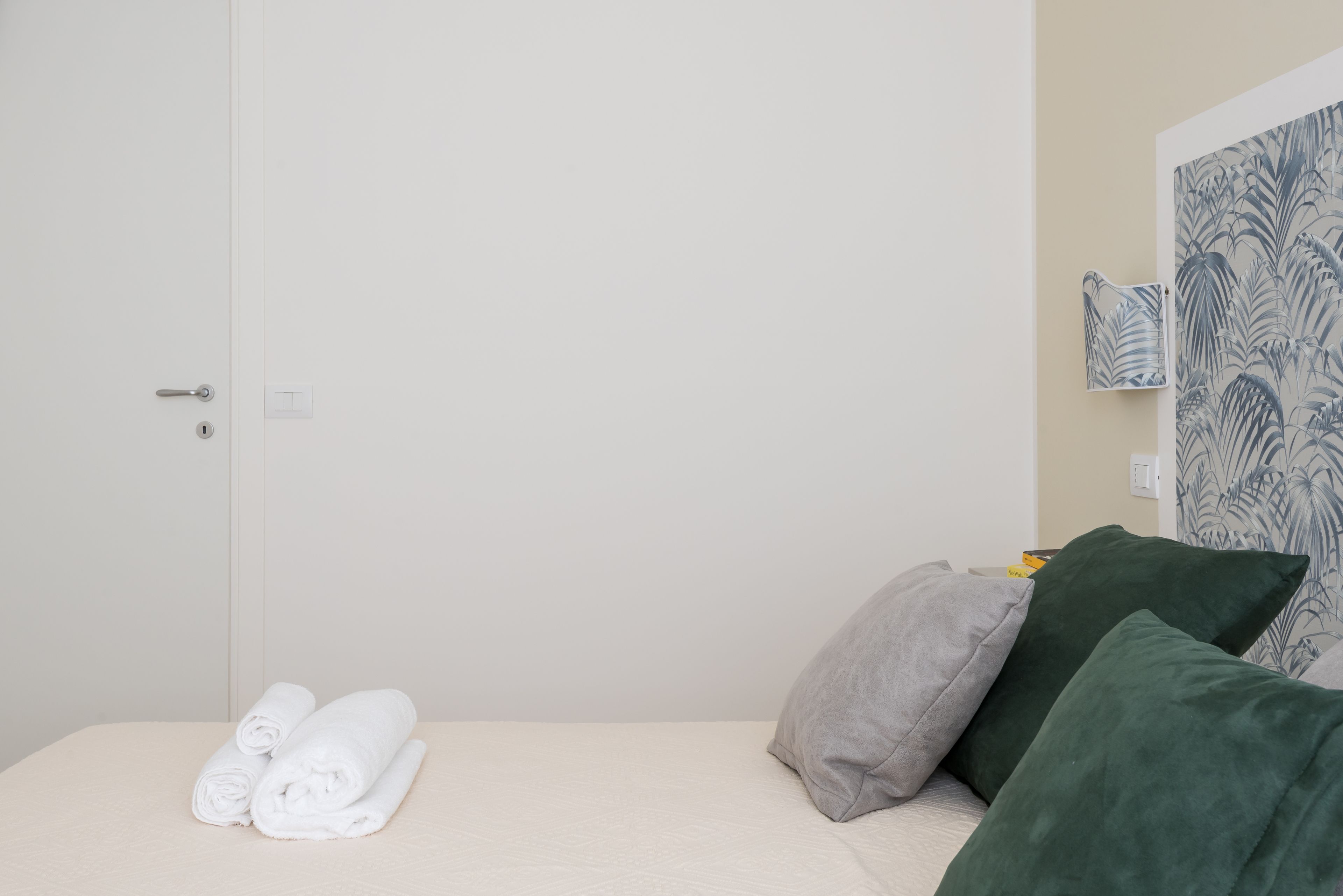 Un lit bien fait, agrémenté de serviettes blanches roulées, une porte blanche et un mur recouvert d’un papier peint tropical aux motifs bleus et blancs.