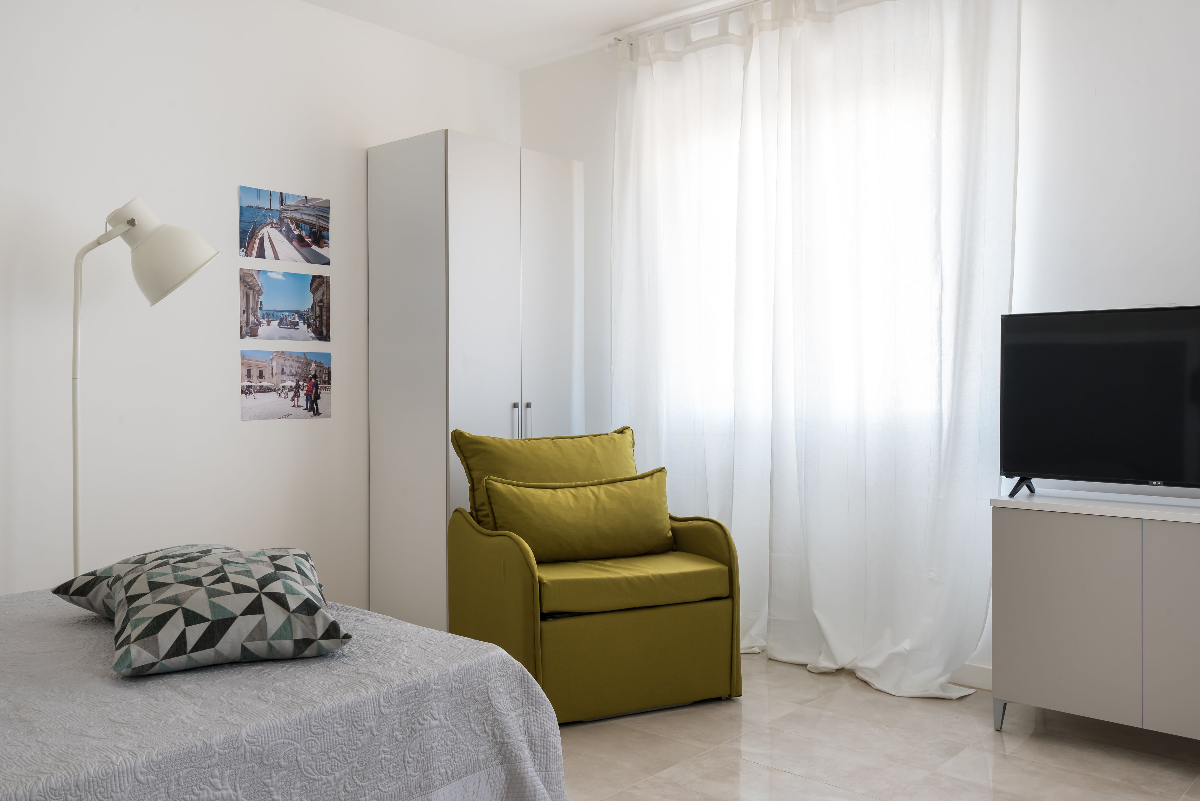 Un dormitorio moderno con una cama, un sillón verde, un televisor sobre un mueble y una pared decorada con fotografías.