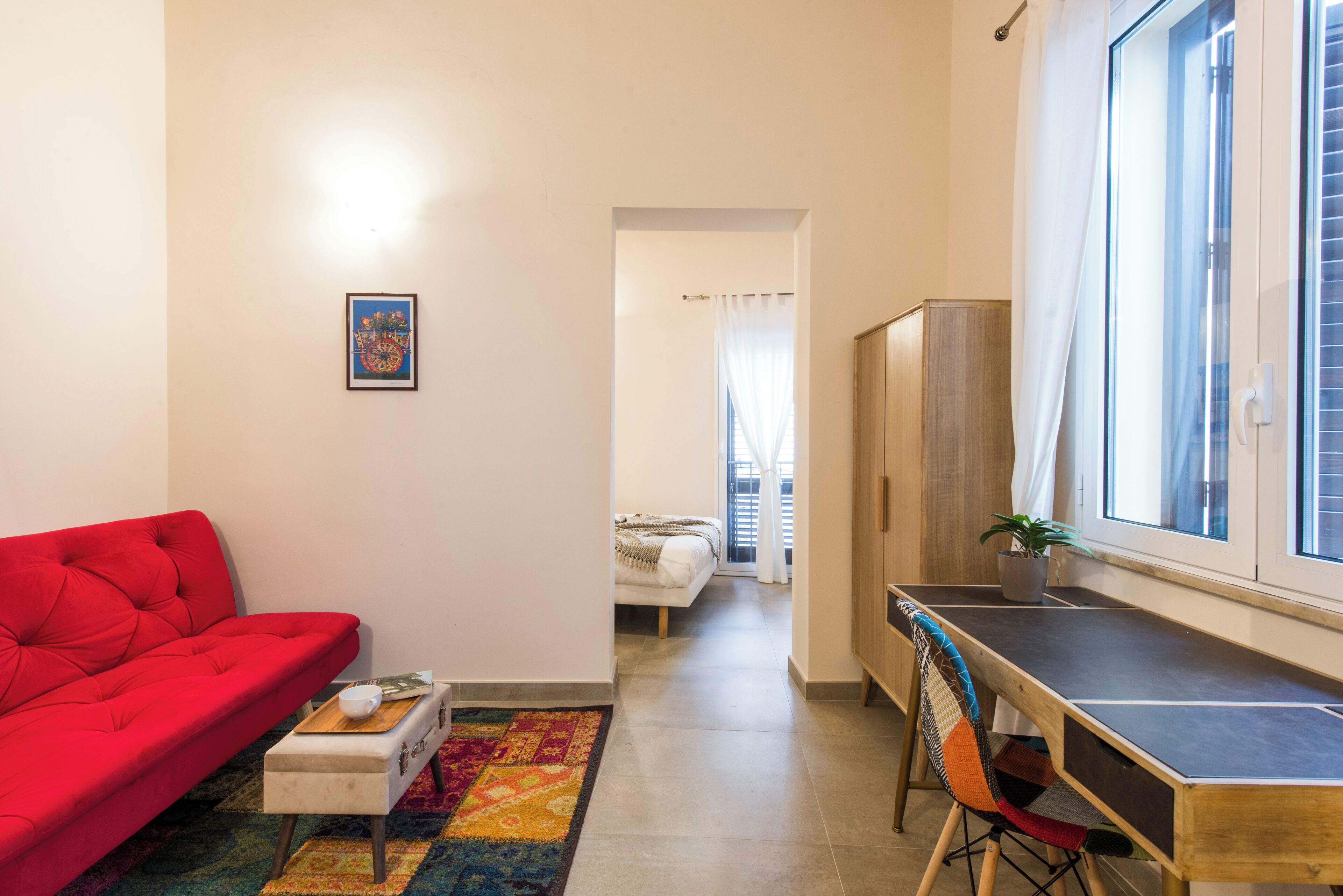 A compact living space with a red sofa, a small table with a cup, a colorful rug, a desk with a chair, and a bed in the background.