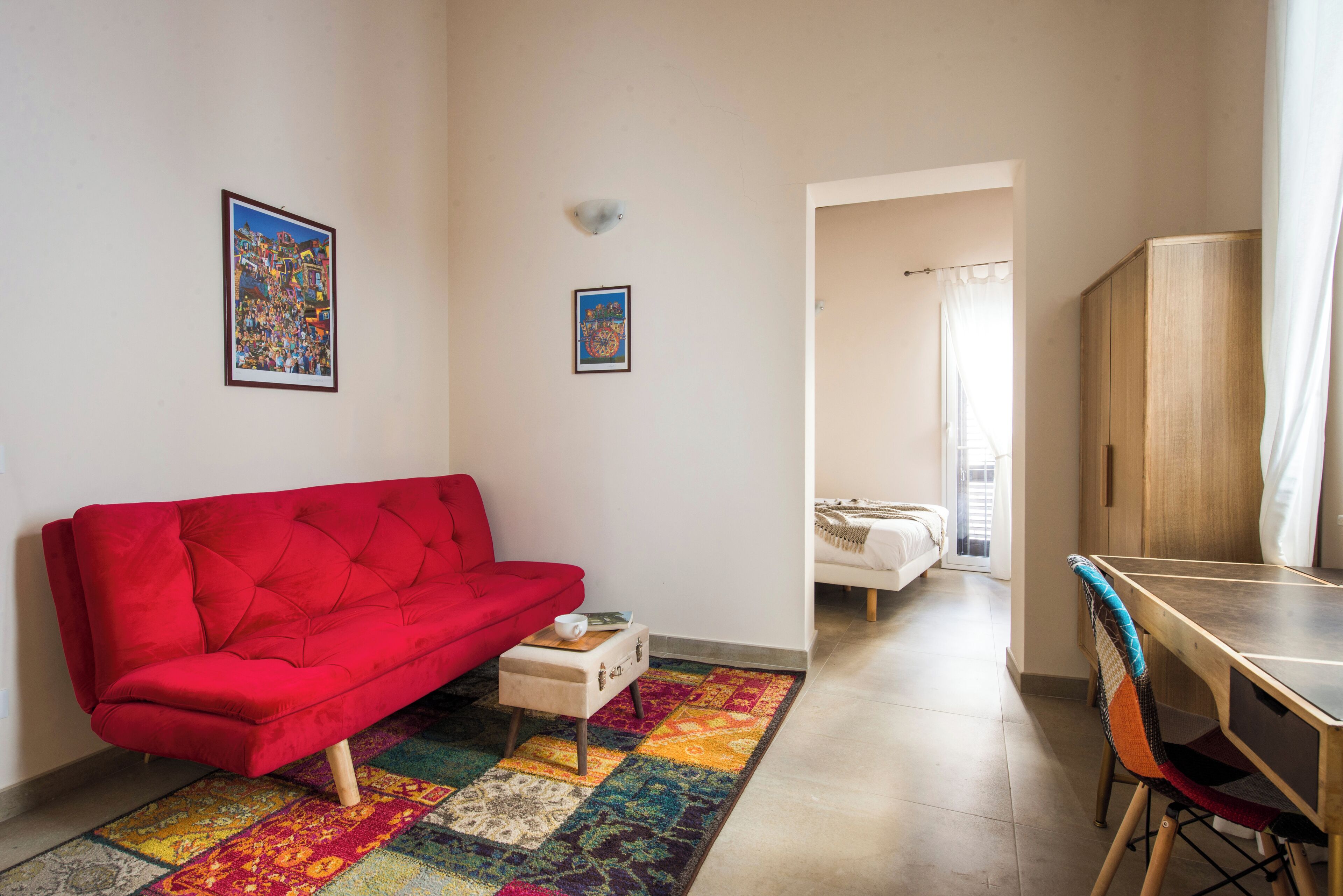 A living room with a red sofa, a small table with a cup, a colorful rug, and a wooden desk with a chair.