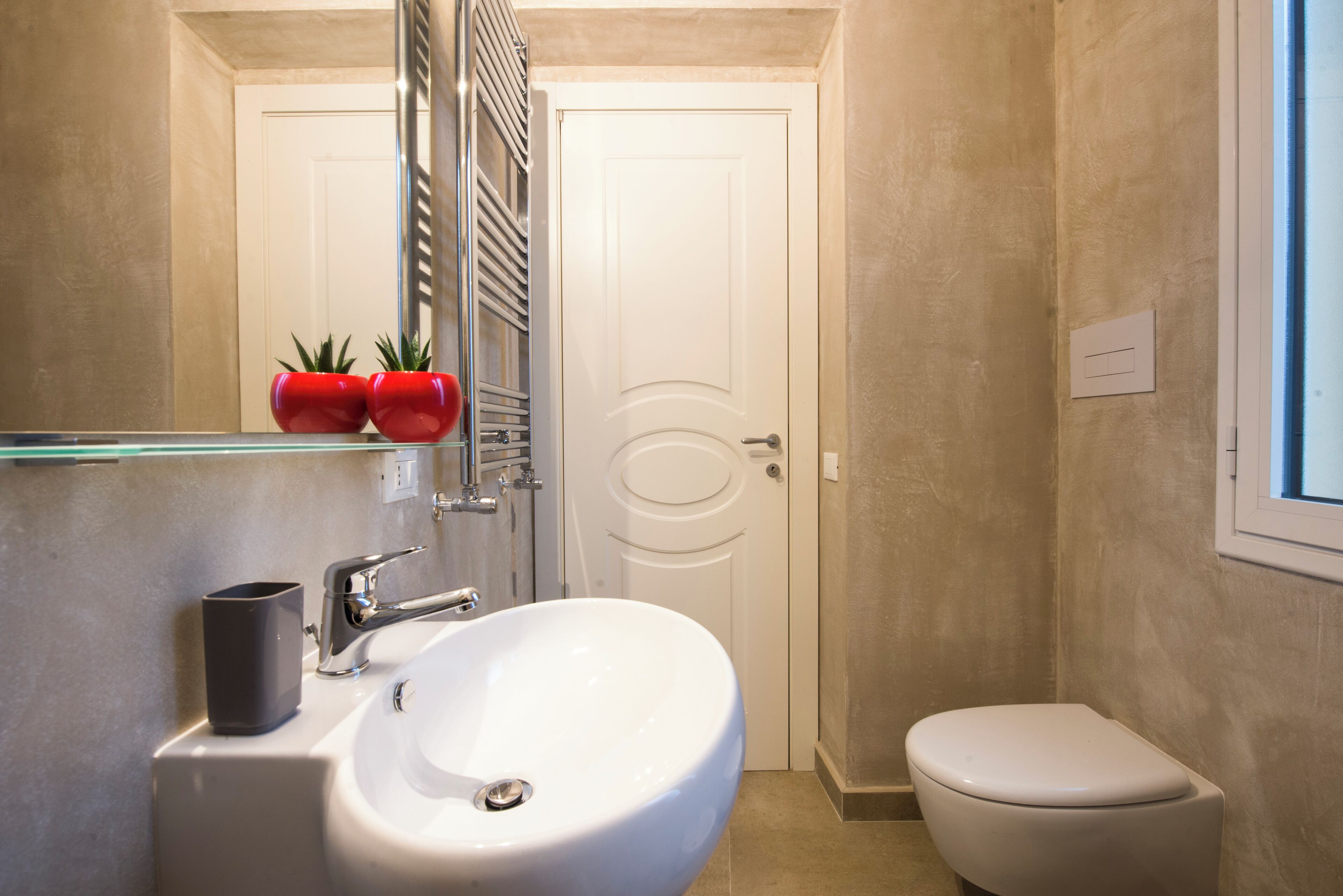 Une salle de bain avec un lavabo blanc, des toilettes et un miroir au-dessus du lavabo. Deux plantes en pot rouges sont posées sur le miroir.