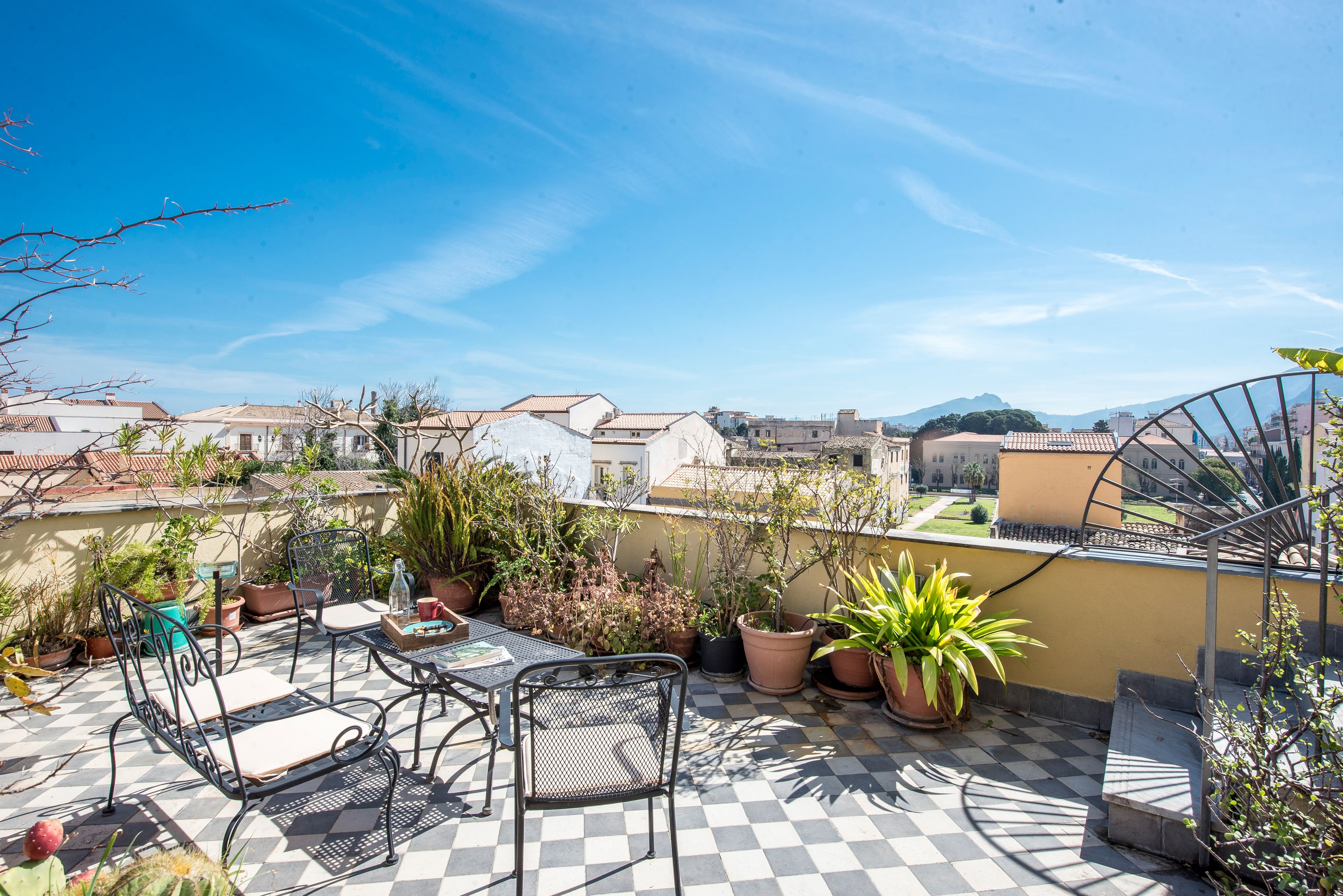 A rooftop terrace with a checkered floor, potted plants, outdoor furniture, and a view of surrounding buildings.