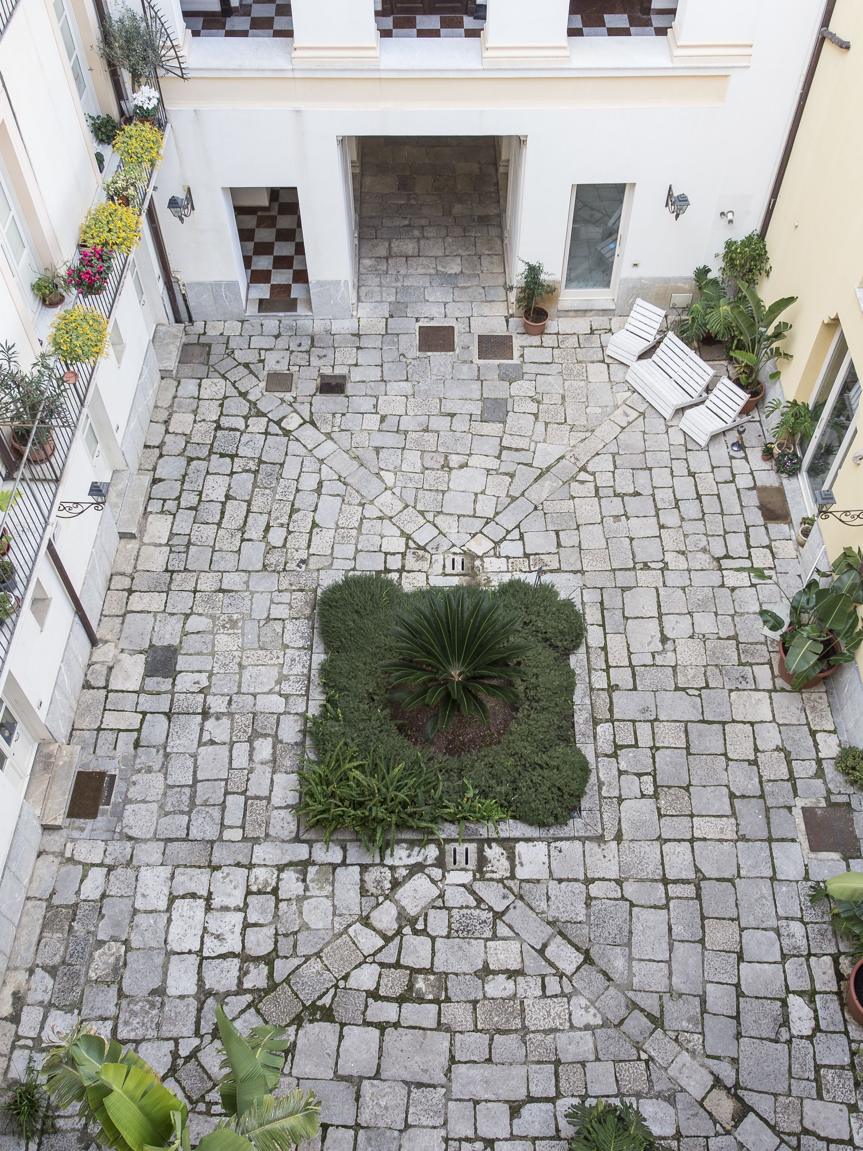 Une cour intérieure avec un jardin central, un sol pavé de pierres et des plantes en pot sur les balcons.