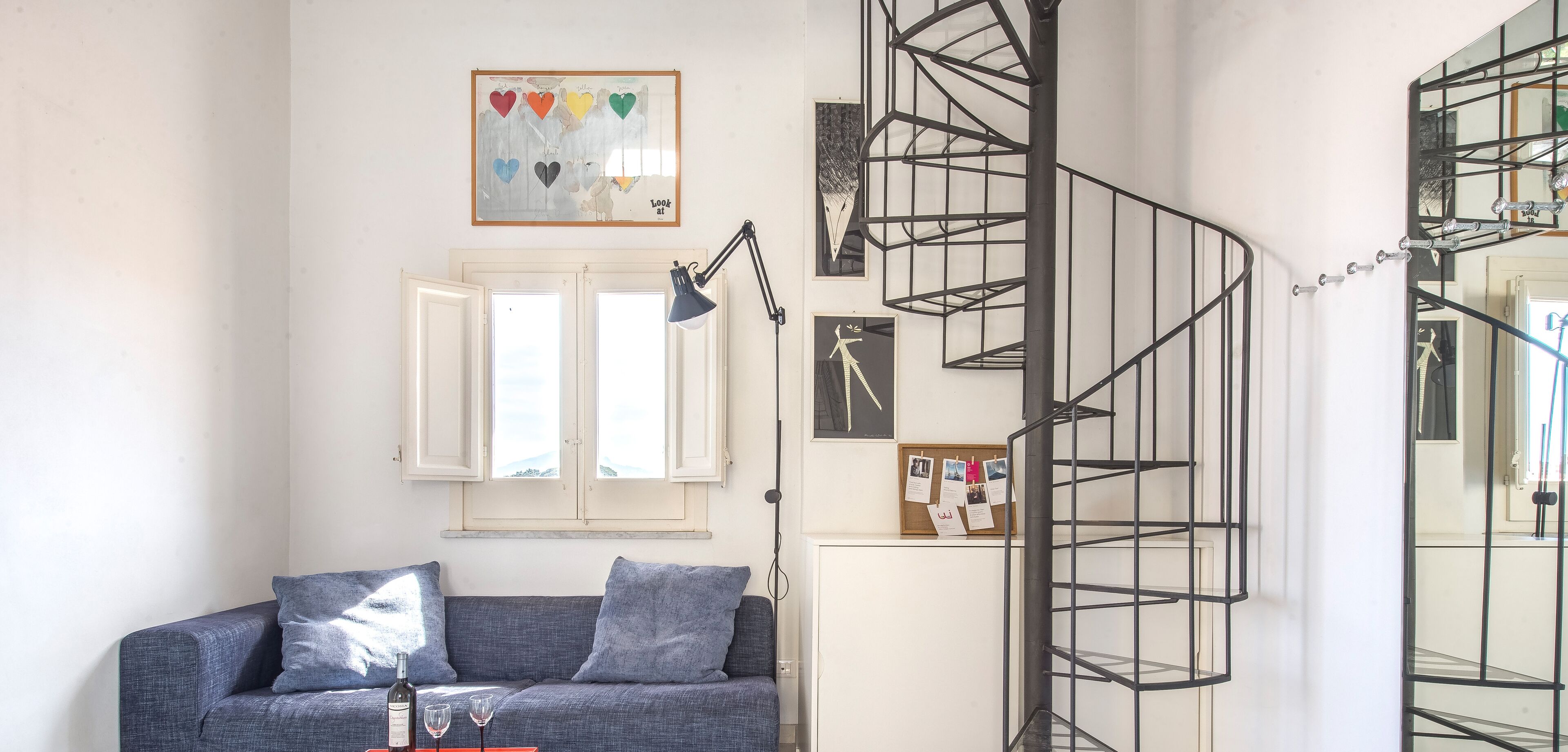Una habitación con un sofá azul, un escritorio con lámpara, una escalera de caracol y un tablón de corcho con fotografías.
