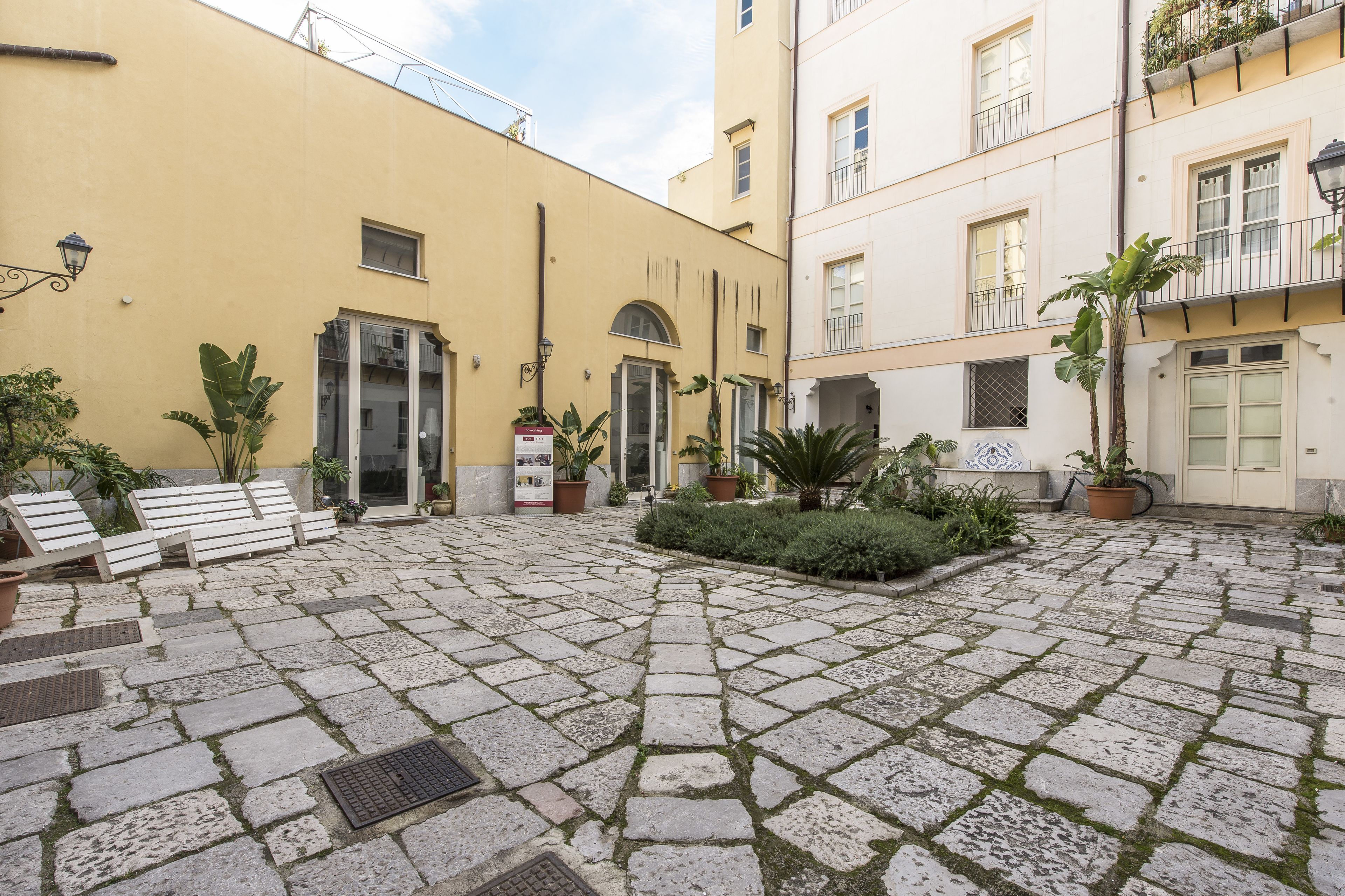 Un cortile con pavimentazione in pietra, panchine, piante in vaso e un edificio con balconi.
