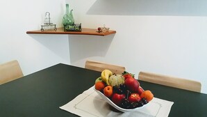 Een eettafel met een fruitschaal in het midden, een houten stoel aan de rechterkant, en een plank erboven met een groene fles en een glas.