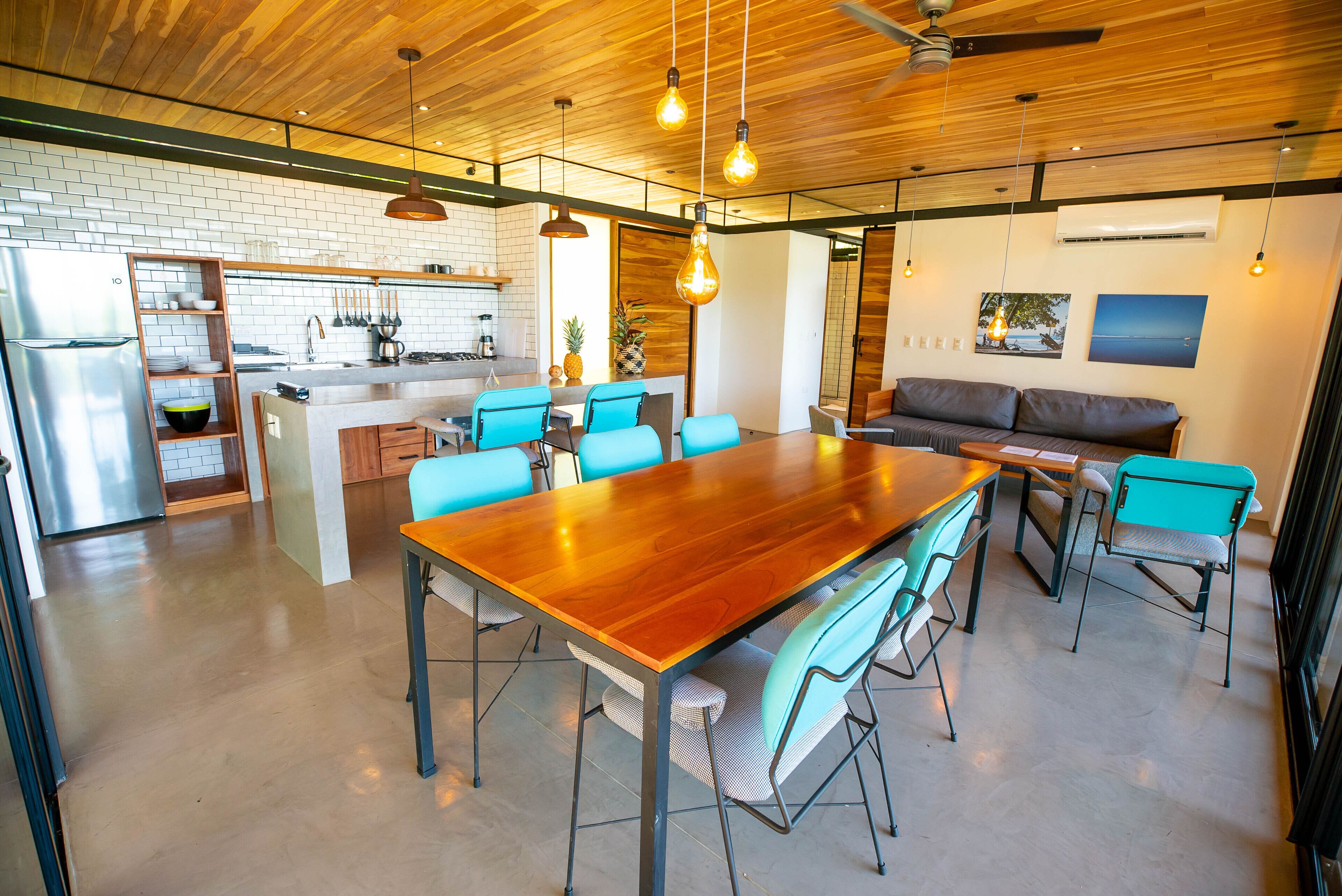 Une cuisine spacieuse avec un îlot central, un coin repas avec une table en bois et des chaises bleues, et un salon avec un canapé et un téléviseur fixé au mur.