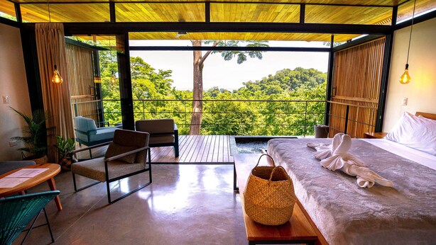 A bedroom with a large bed, a wooden table, a wicker chair, and a view of lush greenery through large windows. - The Green House - Santa Teresa (Cóbano)