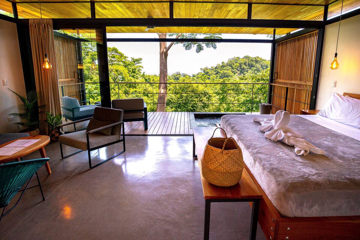 Une chambre avec un grand lit, une table en bois, une chaise en osier et une vue sur une végétation luxuriante grâce à de grandes fenêtres.