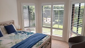 Une chambre à coucher avec un lit, une table de chevet et une chaise. Il y a deux fenêtres équipées de stores et la vue donne sur l’extérieur.