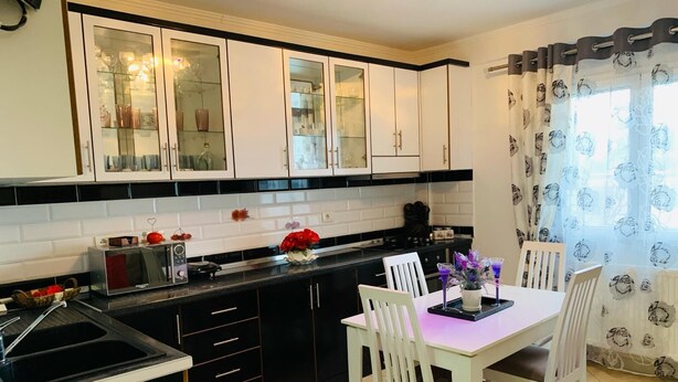A modern kitchen with black and white cabinetry, a dining table with chairs, and a window with floral curtains. - A 3-floor Building With 4 Bedroom, Near Beach (Dërmenas)