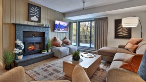 A modern living room with a fireplace, comfortable seating, and a large window offering a view of the outdoors. - The One Everyone Wants in Building 3 at Mountain Green: 1BR, Newly Renovated! (Killington)