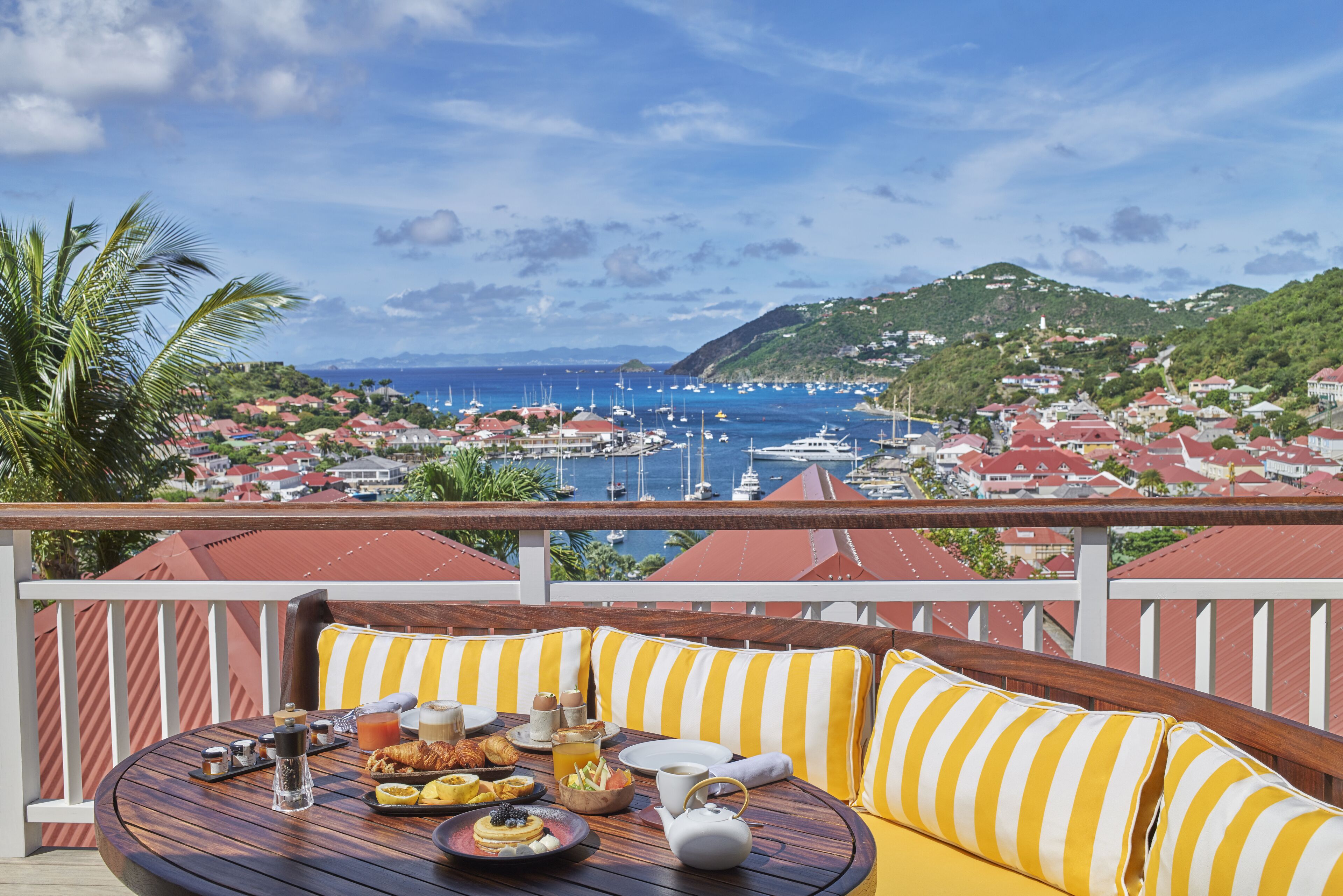 A balcony with a wooden table set for breakfast, overlooking a marina and coastal town.