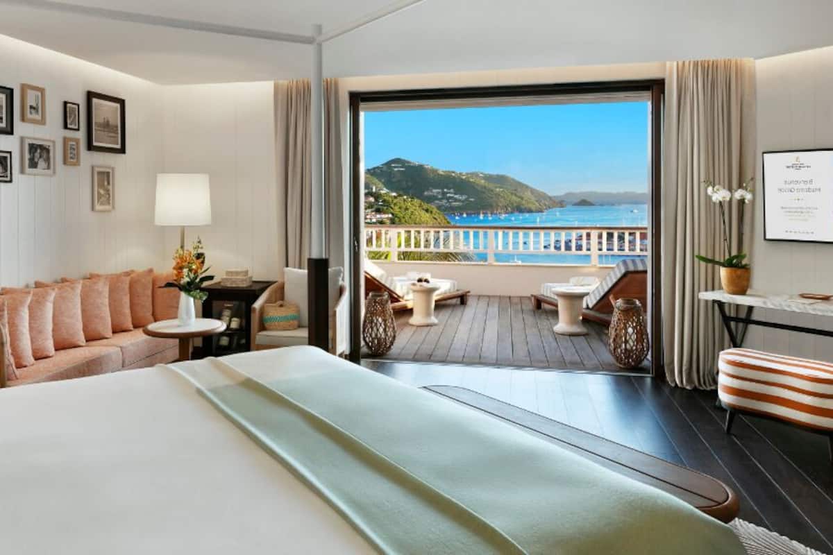 Ein Hotelzimmer mit einem großen Bett, einer Couch, einem kleinen Tisch mit Lampe, einem Balkon mit Meerblick und gerahmten Bildern an der Wand.