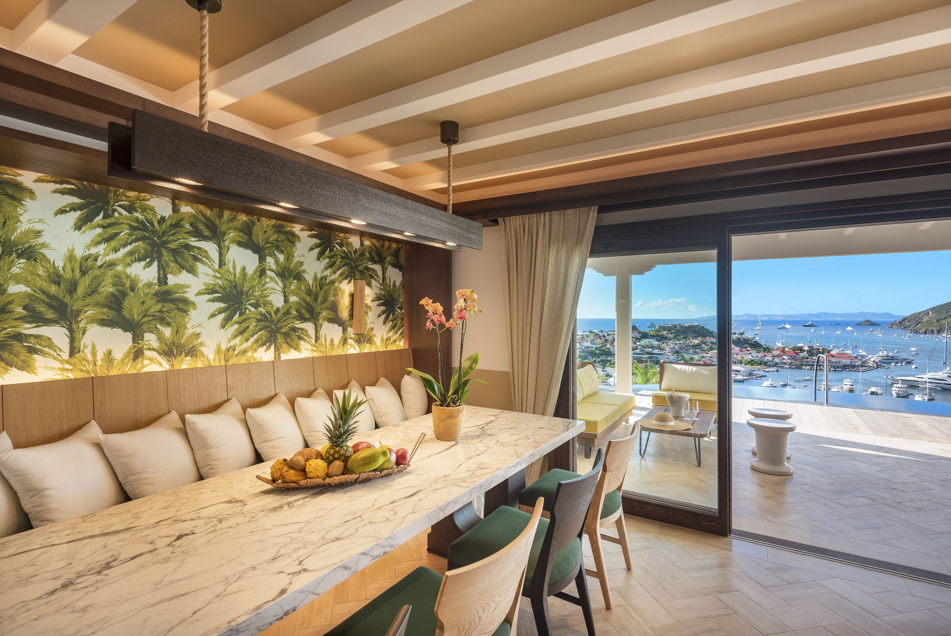 Comedor moderno con una mesa de mármol, sillas con cojines verdes, una cesta de frutas y vistas al mar y a los barcos.