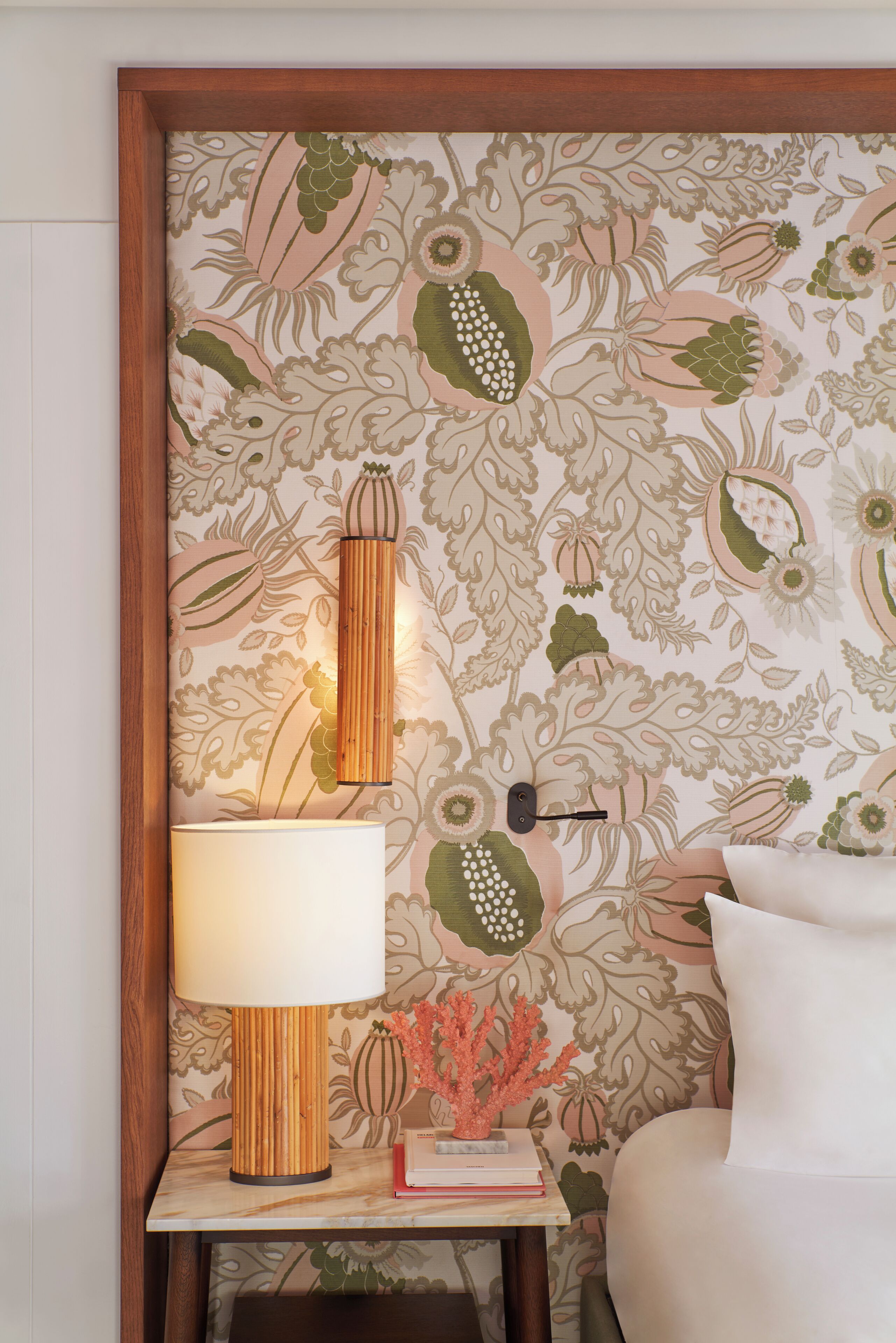 Ein Zimmer mit floralem Wandpapier, einer Nachttischlampe, einem kleinen Tisch mit Büchern und einer Korallen-Dekoration sowie einem weißen Kissen.