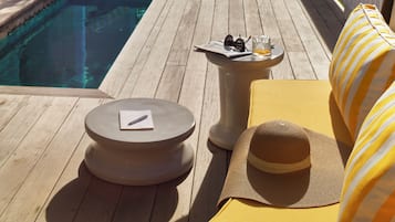 Eine Holzterrasse mit Pool, Liegestuhl, Sonnenliege und einem kleinen Tisch mit Hut und Sonnenbrille.