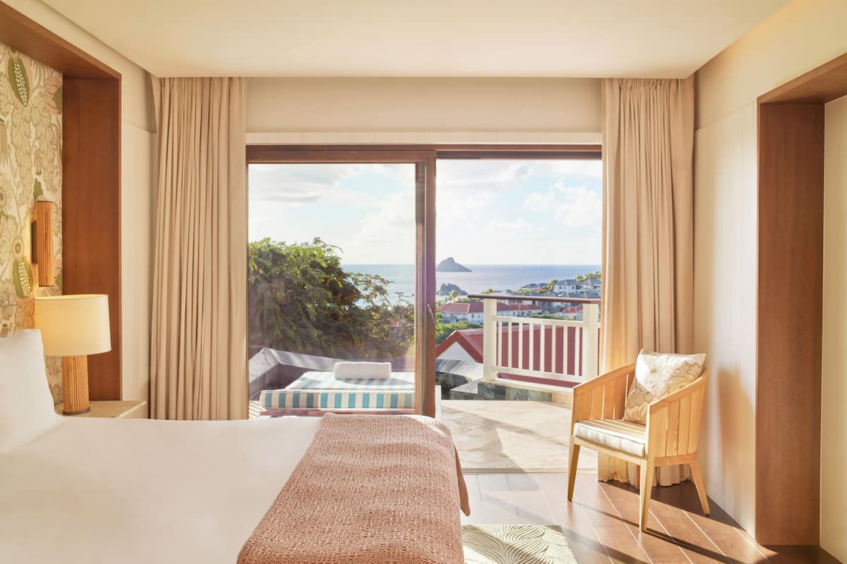 Ein Hotelzimmer mit einem großen Bett, einem Sessel und einem Balkon mit Blick auf das Meer und Gebäude.