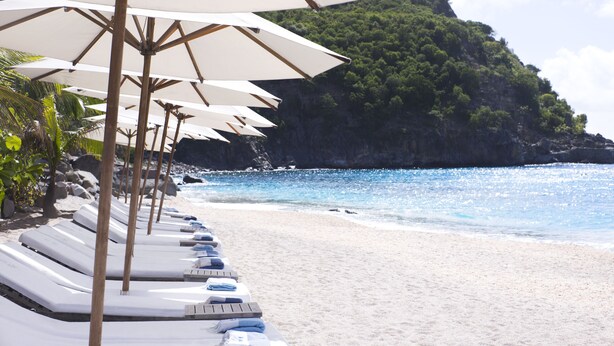 Une plage bordée de rangées de chaises longues sous des parasols, une falaise rocheuse en arrière-plan et une mer bleue et limpide.