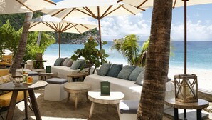 Un salon de plage doté de canapés blancs, de tables rondes et de parasols offrant de l’ombre.