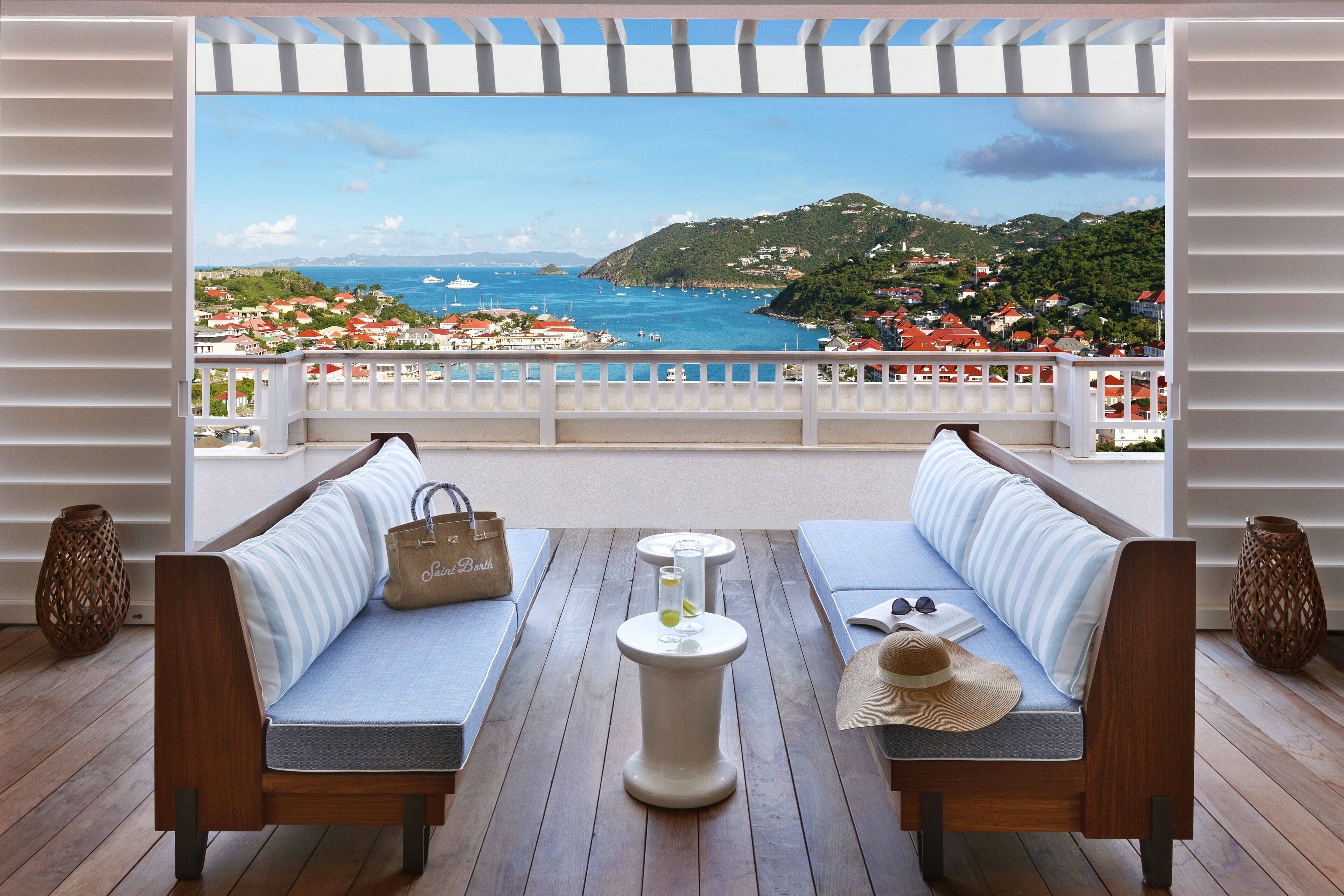 A balcony with a wooden floor, two cushioned sofas, a small round table with a glass and a bottle, and a view of a coastal town.