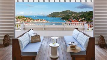 A balcony with a wooden floor, two cushioned sofas, a small round table with a glass and a bottle, and a view of a coastal town.