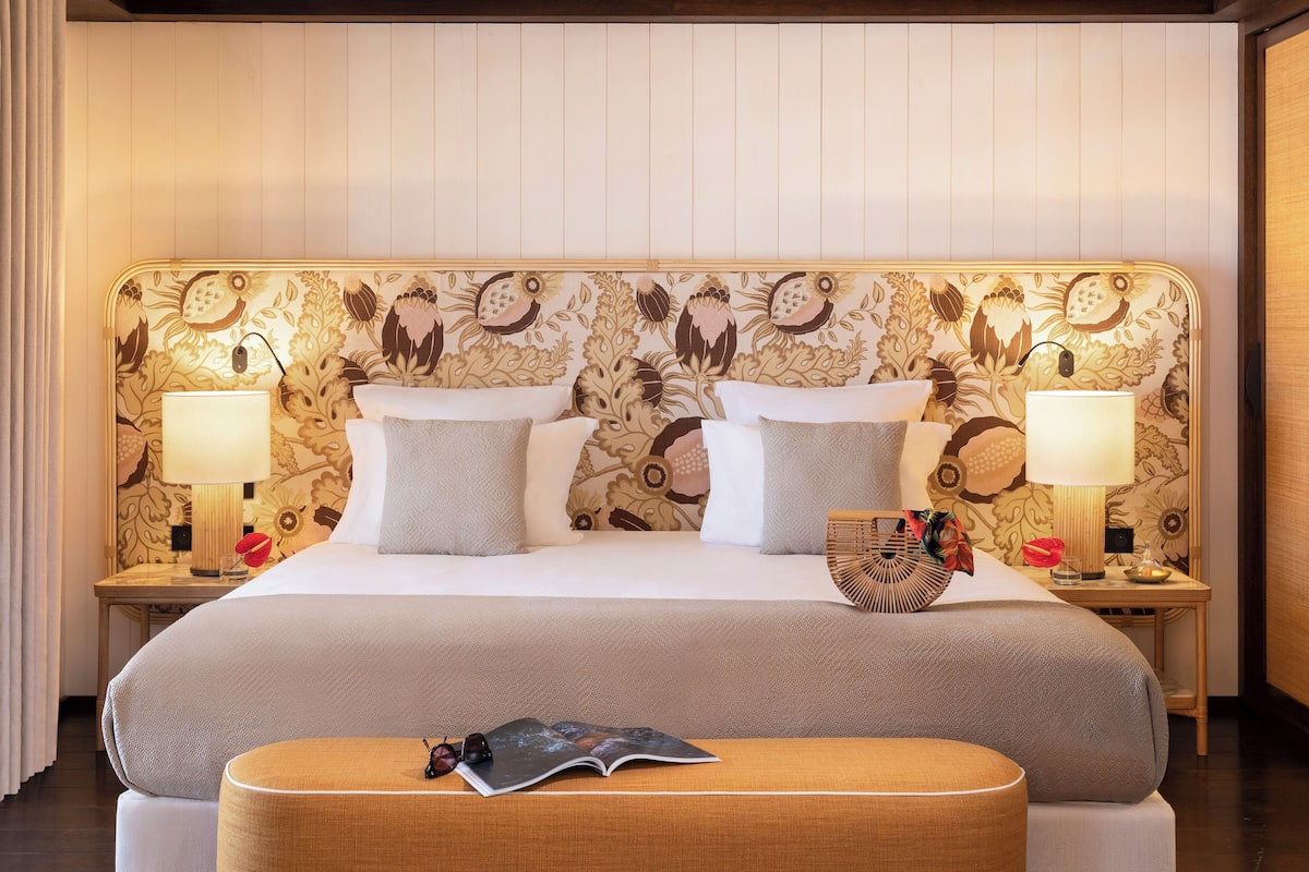 Ein Hotelzimmer mit einem großen Bett, zwei Nachttischen mit Lampen, einem dekorativen Kopfteil und einem Hocker mit einer Zeitschrift.