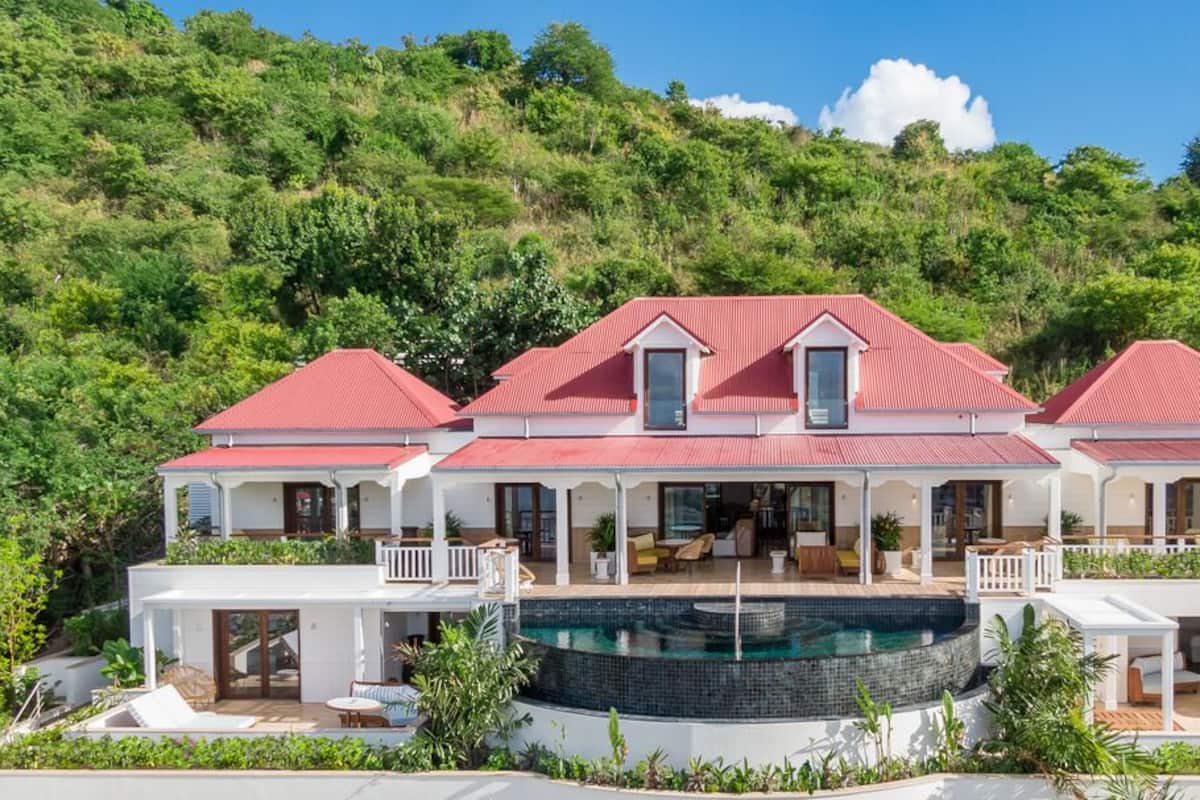 Ein großes, zweistöckiges Haus mit rotem Dach, einem Schwimmbad und einem üppigen, grünen Hang im Hintergrund.