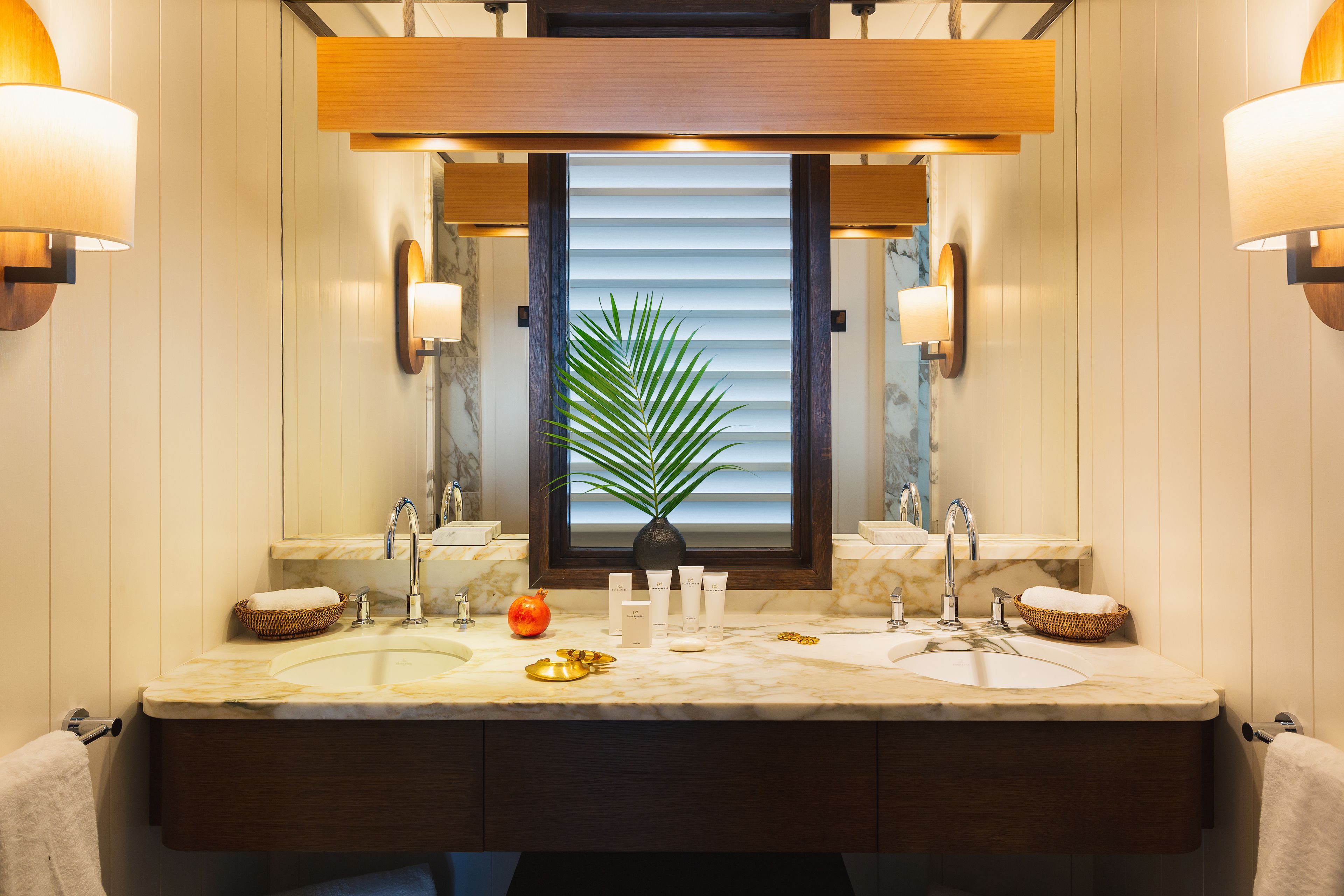 Une salle de bain dotée d’un meuble-lavabo à deux vasques, d’un comptoir en marbre et d’un grand miroir agrémenté d’une plante en pot placée au centre.