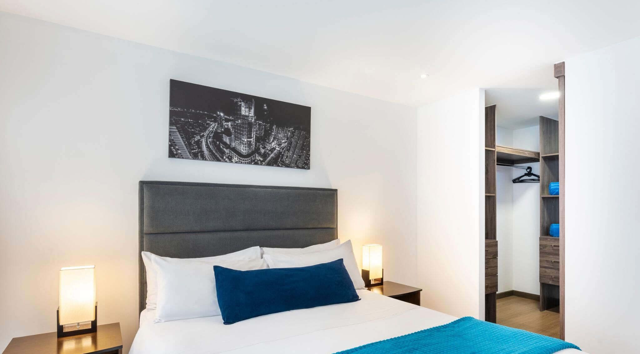 A modern hotel room with a large bed, a cityscape artwork on the wall, and a walk-in closet.