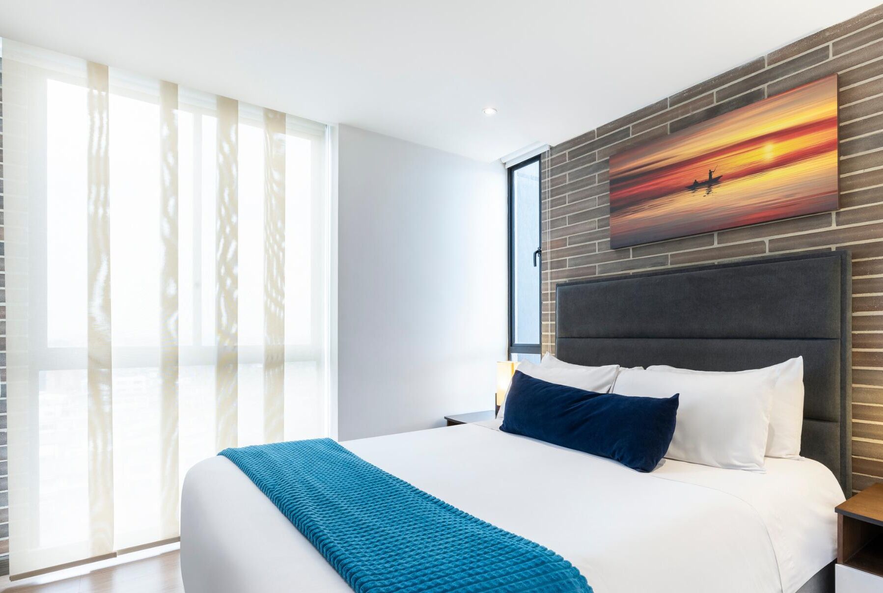 Un hotel moderno con una cama grande, vistas a un atardecer pintoresco y un amplio ventanal con cortinas de gasa.