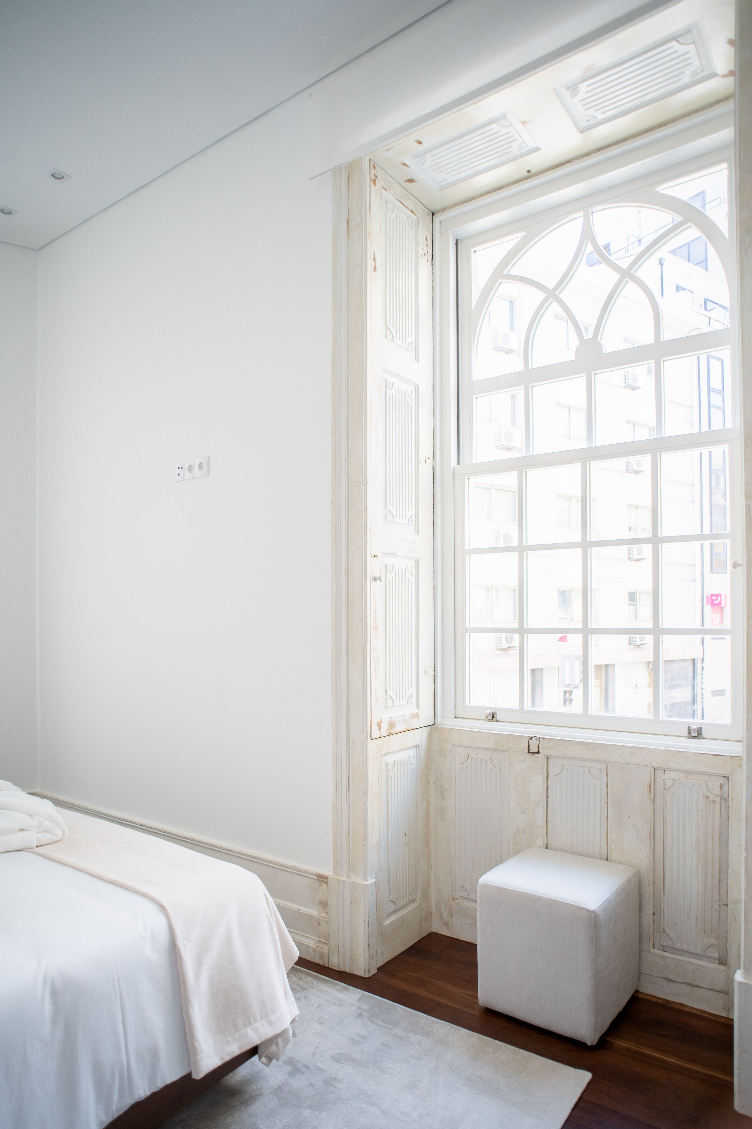 A bedroom with a large window, a bed, a small white ottoman, and a radiator.