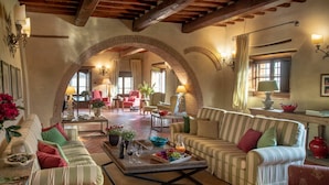 A spacious living area with a high ceiling, exposed wooden beams, and a brick archway leading to another room. There are multiple seating areas with striped sofas and armchairs, a coffee table with a fruit tray, and a fireplace. - Villa Gerlino - Country House with private pool (Rapale)