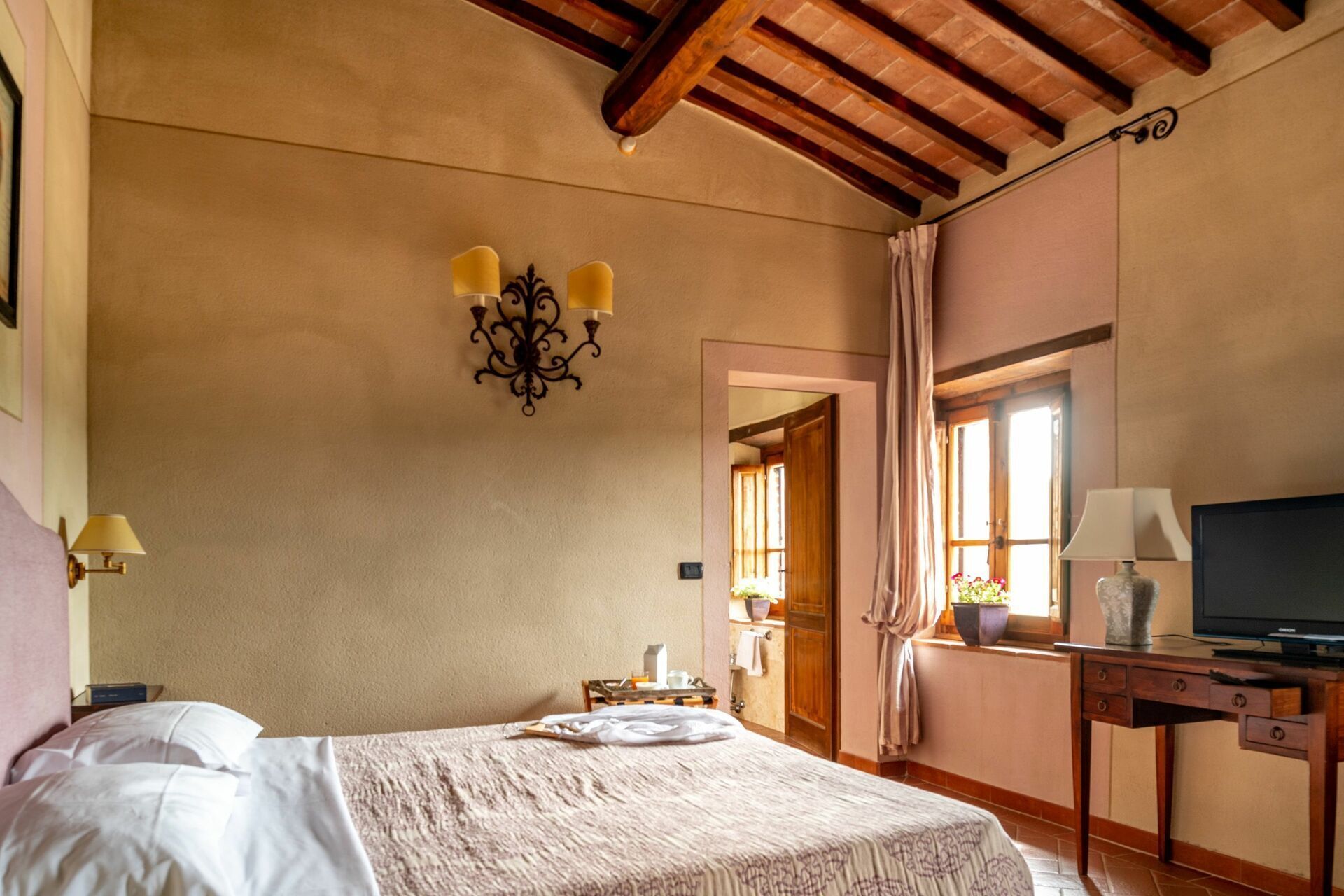 A bedroom with a bed, a desk with a television, a chandelier, and a window with curtains.