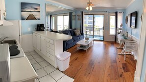 A compact kitchen with a sink, stove, and white cabinets. A blue sofa and a small table with a decorative item are in the living area. - Tybee Island Ocean Therapy (Tybee Island)