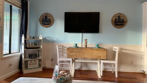 A dining area with a wooden table and white chairs, a TV mounted on the wall, and decorative wall art. - Tybee Island Ocean Therapy (Tybee Island)
