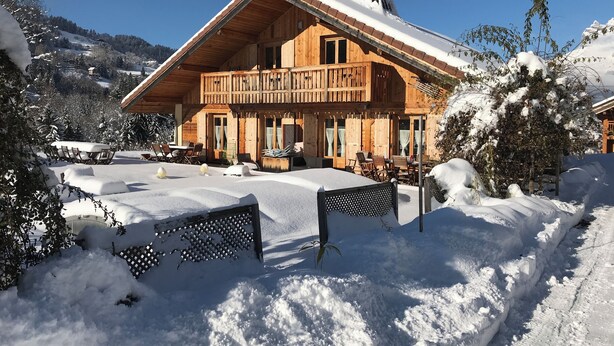 A wooden chalet with a balcony, surrounded by snow-covered trees and a clear sky.