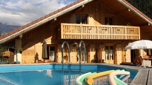 A wooden chalet with a pool, a balcony, and a mountain backdrop.