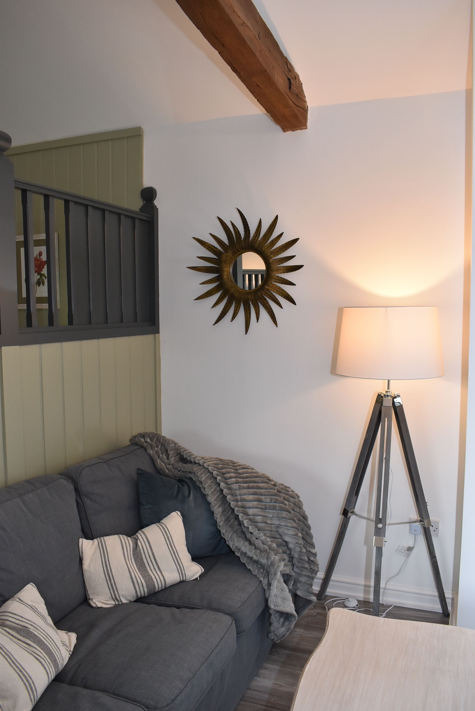 Ein Zimmer mit einer grauen Couch, einem gestreiften Kissen, einer Wanddekoration in Sonnenform, einer Stehlampe und einer Treppe mit Holzgeländer.