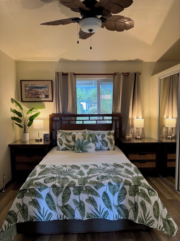 A bedroom with a bed, a ceiling fan, a window with curtains, a nightstand with a lamp, and a framed picture on the wall.