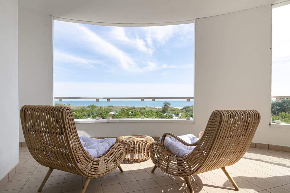 A balcony with two wicker chairs and a small table, overlooking a beach and ocean.