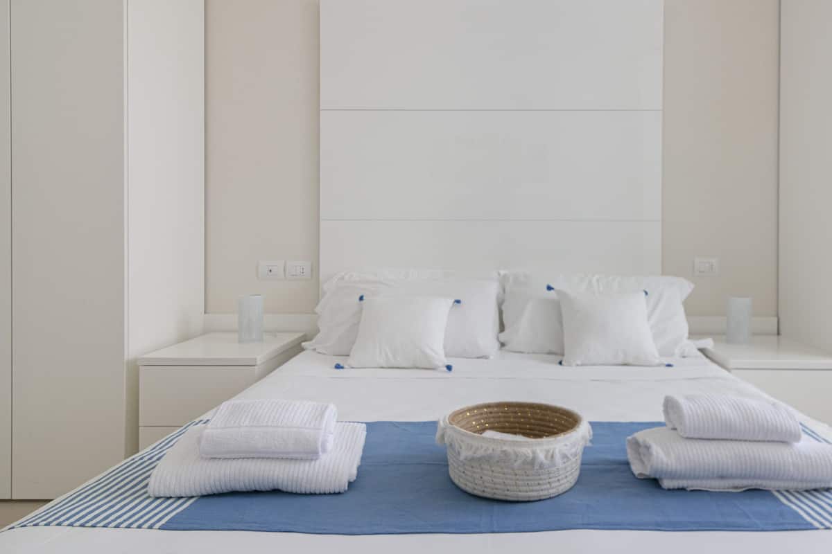 A hotel room with a bed, white pillows, a blue bedspread, and a woven basket.