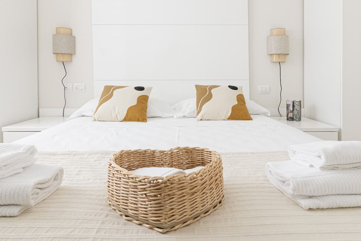 A hotel room with a neatly made bed, two bedside lamps, a wicker basket, and folded towels on the floor.