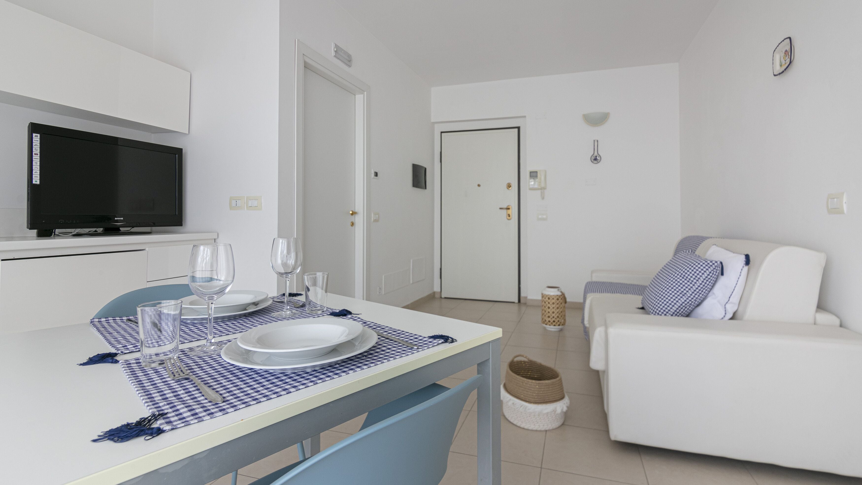 En moderne, veloplyst stue med en hvid sofa, et spisebord dækket med tallerkener og glas, et fjernsyn og en dør der fører til et andet rum.