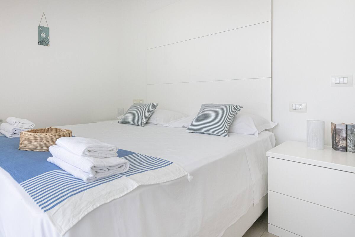 A neatly made bed with white and blue striped bedding, a basket of folded towels, and a bedside table with a lamp and books.