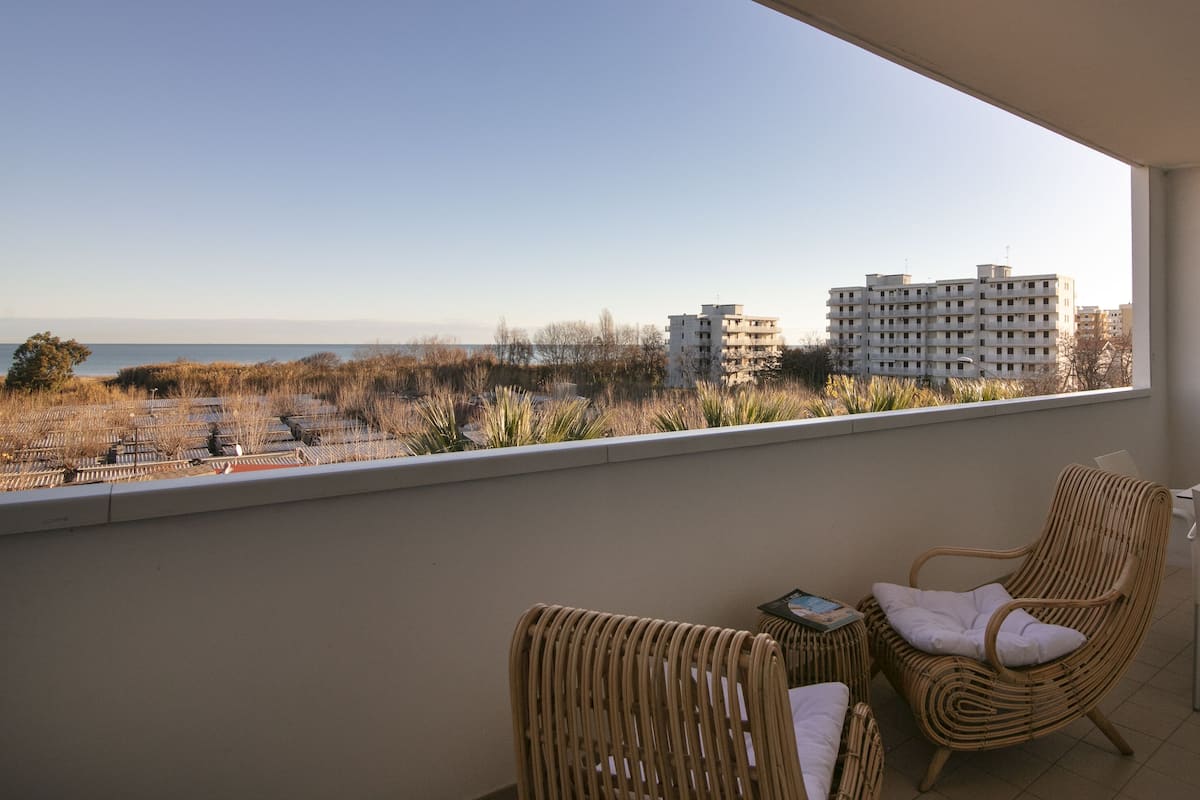 A balcony with wicker chairs and a table, overlooking a cityscape and the sea.