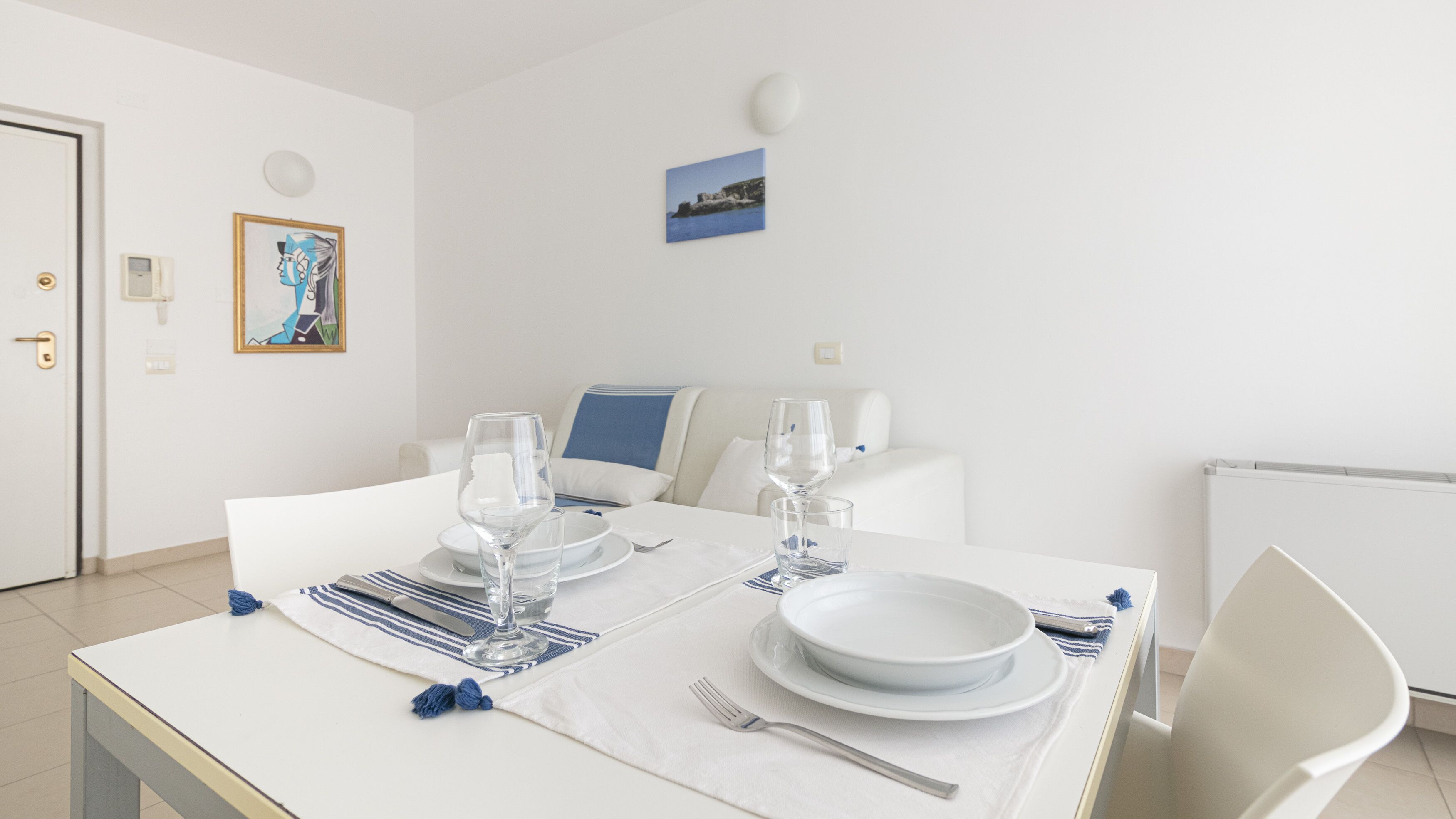A dining area with a table set for two, a white sofa, and a framed picture on the wall.